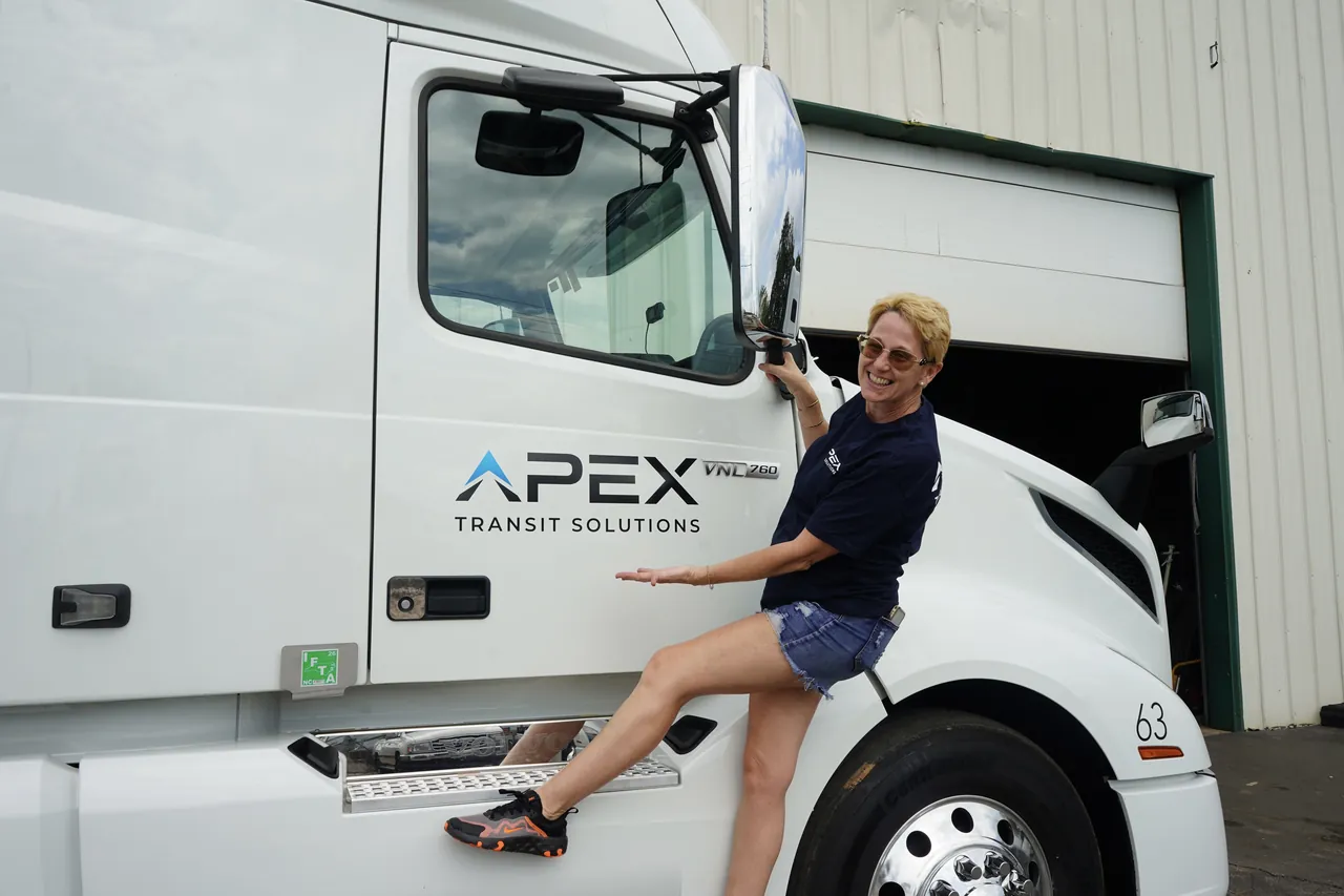 A smiling team member poses on the step of a white Apex Transit tractor, next to an open warehouse bay door.
