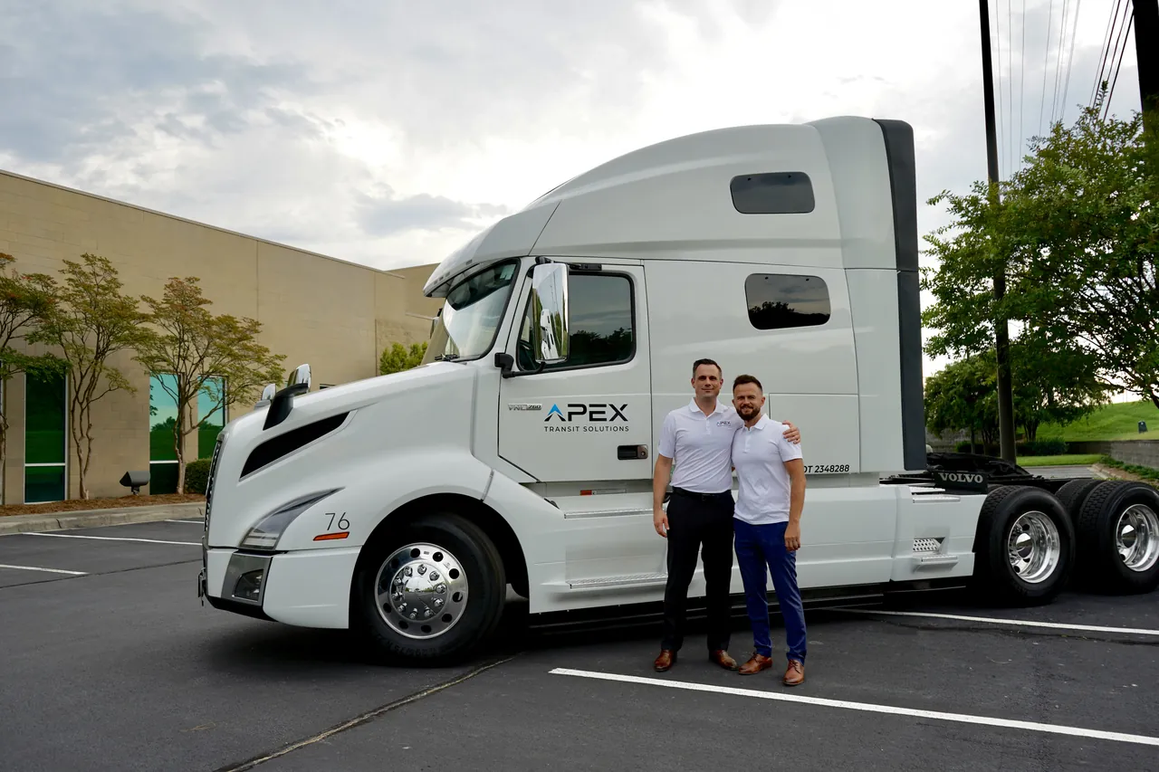 Two team members in white Apex polo shirts stand together in front of a white Apex Transit tractor in an office parking lot.