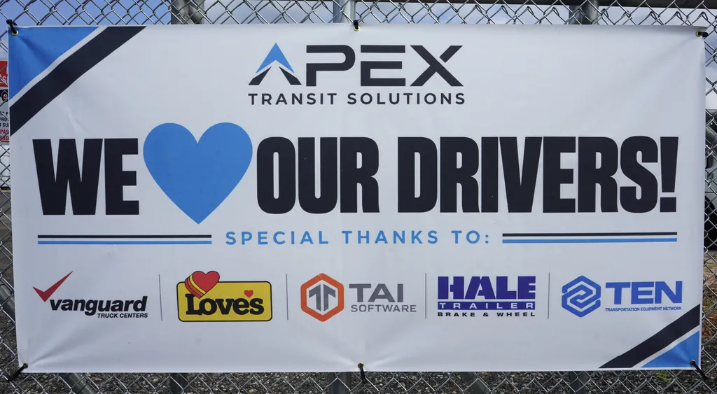 A banner on a chain link fence reads: Apex Transit Solutions. We love our drivers! Special thanks to Vanguard Truck Centers, Love's, TAI Software, Hale Trailer Brake and Wheel, and TEN Transportation Equipment Network.