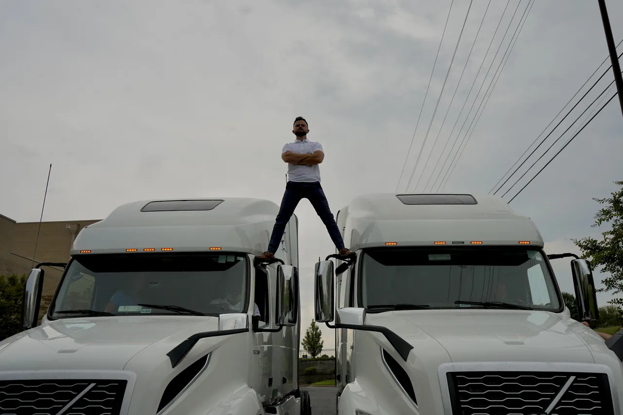 A team member stands with arms crossed, balanced across the gap between two white tractors parked side by side under an overcast sky.