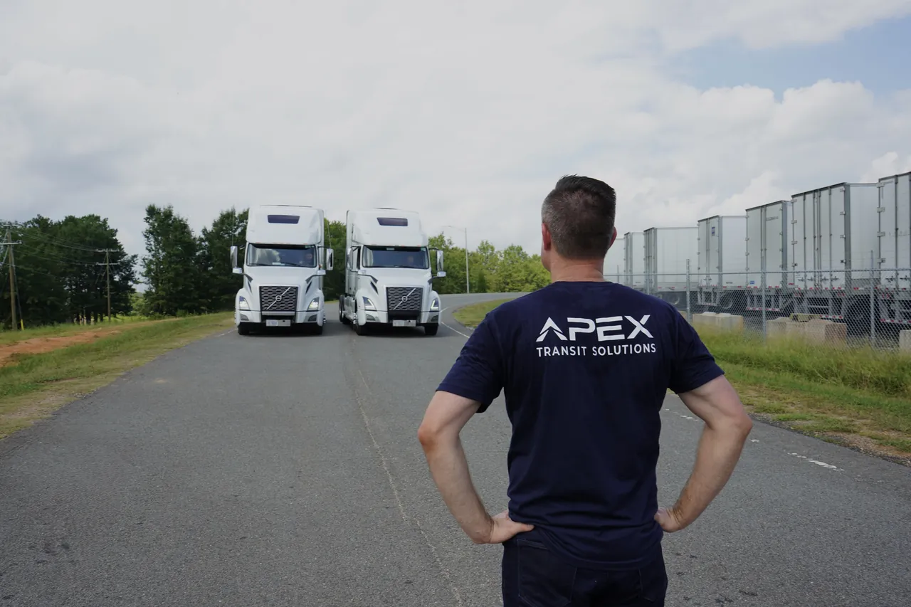 A team member in an Apex Transit Solutions shirt, seen from behind, faces two white Apex tractors parked side by side on a road, with a row of trailers to the right.