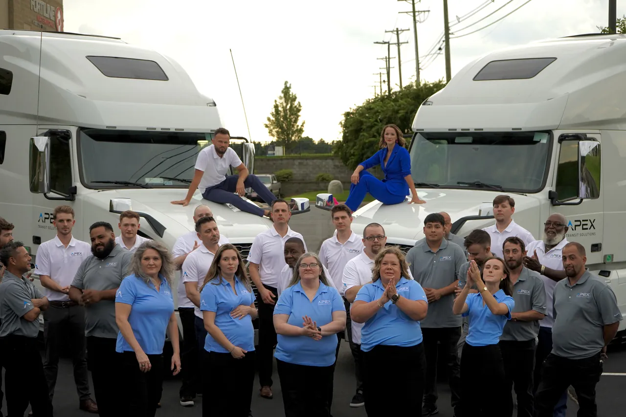 A large group of Apex Transit team members in Apex polo shirts, gathered in a parking lot in front of two white Apex trucks, with two team members seated on the truck hoods.